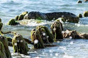 Broken wooden pier remains in sea. Beautiful water color under sunlight. Tide and sea spray. Old wooden posts overgrown seaweed. photo