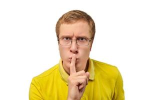 Funny young man in yellow T-shirt asking to be quiet, silence gesture, white background photo