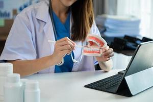 dentista sentado a la mesa con muestras de mandíbula modelo de diente y trabajando con tableta y computadora portátil en el consultorio dental clínica dental profesional. medico trabajando foto