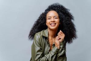 Joyful woman with dark skin, Afro hairstyle, keeps hands together, smiles sincerely at camera, shows white teeth, wears leather shirt, poses over grey background. People, emotions, ethnicity photo