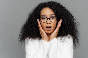 Omg, it cannot be so Surprised emotional dark skinned young female with curly hair, touches both cheeks, has shocked faciall expression, dressed in white jumper, isolated over grey background photo