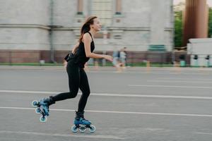 concepto de patinaje y fitness de verano. Una mujer joven de cabello oscuro en forma, aficionada a los ejercicios de patinaje sobre ruedas, todo el cuerpo posa en la carretera a lo largo del asfalto durante el día soleado, usa ropa cómoda y tiene una figura deportiva foto