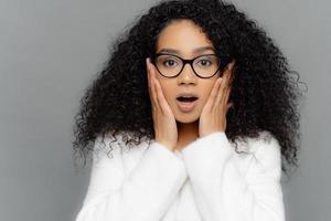 Close up portrait of shocked concerned young woman with dark skin and curly hair, hears terrifying news, keeps hands on cheeks, wears spectacles and white sweater, isolated on grey background photo