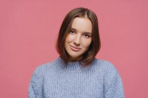 Close up shot of satisfied dark haired young lady tilts head, enjoys lovely casual talk, wears warm winter sweater, poses alone against pink background, gazes focused at camera, feels relaxed photo