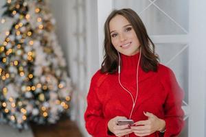 Cheerful good looking woman has toothy smile, enjoys nice track in earphones, updates playlist on smart phone, dressed in warm knitted sweater, poses against decorated Christmas tree in background photo