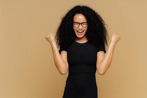 foto de estudio de una mujer alegre y alegre con el pelo rizado, levanta los puños apretados, celebra el éxito y el triunfo, vestida con una camiseta negra informal, anteojos, se para contra una pared marrón con un espacio vacío