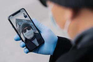 Shot of man has video conference via modern smartphone, being on distance from people during coronavirus spreading, wears protective mask and rubber gloves while walks outdoor in public place photo