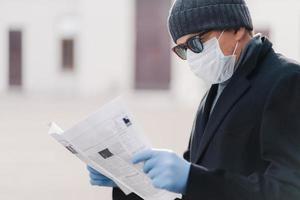 Quarantine for coronavirus. Horizontal shot of adult man wears protective mask and disposable gloves, reads fresh newspaper, finds out news about pandemic situation. Infection prevention concept