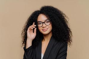 Close up portrait of calm satisfied Afro American woman with bushy curly hairstyle, keeps hand raised, has healthy skin, wears spectacles, black elegant outfit isolated on brown wall. Face expressions photo
