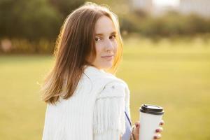 Relaxed attractive young female model with dark hair, drinks takeaway coffee, stands back, has outdoor stroll, enjoys sunny warm day, looks directly at camera. People, rest and lifestyle concept