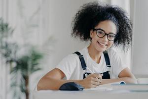 Time for studying. Happy dark skinned teenage girl holds pen, writes down information needed for course work, busy preparing for classes, dressed in casual wear and spectacles, being hard working photo