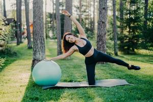 Horizontal shot of active slim woman with cheerful expression leans aside does exercises with fitness ball, dressed in sportsclothes, poses outdoor against tall trees on karemat. Healthy lifestyle photo