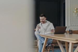 Joyful young male entrepreneur sitting near wooden table and using smartphone while working remotely on laptop from home photo