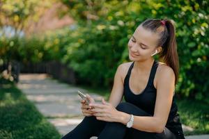 Pleased active woman with pony tail wears sportive wear concentrated at smartphone device listens music in wireless earphones browses internet has outdoor training. Healthy lifestyle concept photo