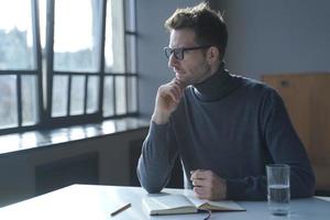 Young pensive thoughtful German man sitting at desk and looking out window photo