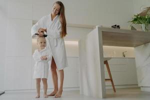 Loving mom brushing hair of cute little daughter after taking bath at home photo