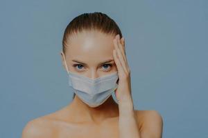 Covid 19 concept. Serious brunette European woman touches face looks confidently at camera has combed hair makeup wears medical protective mask during virus pandemic stands shirtless indoor. photo