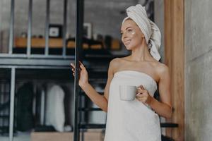 Pretty thoughtful woman concentrated somewhere, smiles broadly, has white perfect teeth, applies face cream, stands wrapped in towel, drinks hot tea, stands on stairs indoor, relaxes after taking bath photo