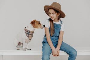 Isolated shot of pretty girl wears stylish hat, white t shirt and denim overalls, looks at her puppy, plays with pet, loves animals, being true friends, pose on table, isolated on white background photo