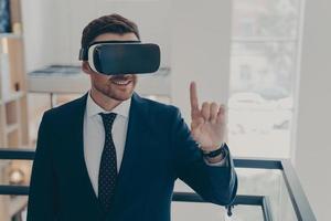 Smiling successful businessman in suit standing in office interior and using VR glasses for work photo