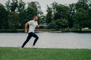 Horizontal shot of handsome active bearded man goes running being photographed in motion, runs against river and beautiful nature, gets ready for marathon. Sport, wellness and workout concept photo