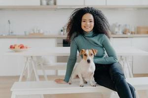 Happy housewife with Afro haircut, sits at bench with pedigree dog, have fun and look directly at camera, pose in kitchen, express good emotions, relax together. Lovely pet with owner at home photo