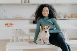 ama de casa feliz con corte de pelo afro, se sienta en el banco con un perro pedigrí, diviértete y mira directamente a la cámara, posa en la cocina, expresa buenas emociones, relájate juntos. adorable mascota con dueño en casa foto