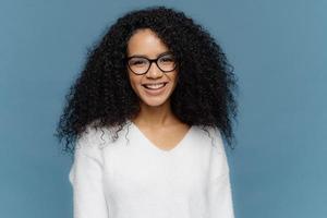 Headshot of lovely curly woman being in good mood, smiles broadly, expresses nice feelings, wears transparent spectacles and white jumper, isolated over blue background. Afro female poses indoor photo