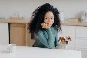 Photo of pleasant looking woman dressed in casual wear, has curly bushy hair, poses against kitchen interior with pedigree dog, has morning coffee, enjoys weekend, smiles pleasantly at camera