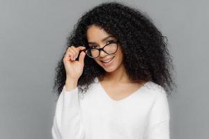 Horizontal shot of satisfied female teacher keeps hand on frame of glasses, smiles happily, has Afro hairstyle, wears casual white jumper, stands indoor over grey background. Good mood and beauty photo