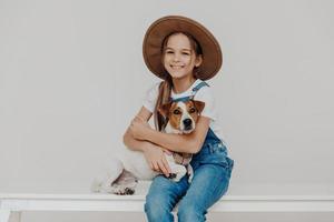 Indoor shot of adorable small preschool girl dressed in fashionable outfit, huggs jack russel terrier dog, plays with best animal friend, love each other. Children, pets and friendship concept photo