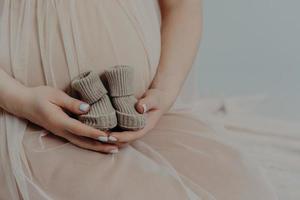 concepto de maternidad y embarazo. la mujer embarazada irreconocible sostiene botas pequeñas sobre la barriga y viste un vestido festivo. futura madre sin rostro foto