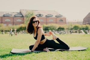 Sports activity concept. Horizontal shot of happy motivated woman stretches on fitness mat trains outdoors during summer day has sporty healthy body wears sunglasses cropped top and leggings photo