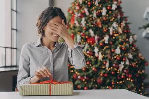 Excited european woman getting christmas surprise, sitting at desk near decorated xmas tree at home photo