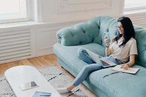 Above shot of pretty brunette woman focused in samrtphone display, writes down necessary information in notebook, watches training webinar, poses in cozy room on comfortable couch, works from home photo