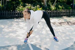 Horizontal shot of dark haired athletic European woman involved in workout routine leans to foot exercises outdoor wears activewear smiles positively warms up trains muscles poses full length photo