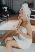 Vertical shot of relaxed happy woman applies moisturising cream on face, cares about complexion and skin, sits over domestic atmosphere, wrapped in bath towel. Facial treatment and hygiene concept photo