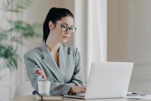 Serious female entrepreneur in eyeglasses, has dark hair combed in pony tail, works on business project, concentrated at laptop computer wears grey formal costume uses information from web resources photo
