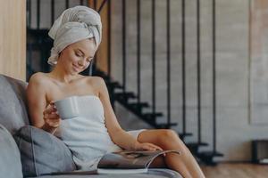 Indoor shot of relaxed beautiful woman wrapped in towel, drinks aromatic coffee from cup, sits on comfortable sofa in living room, reads magazine, spends free time at home. Domestic atmosphere photo