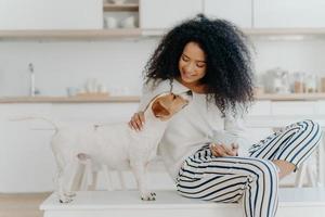 Delighted curly woman with cheerful expression poses with jack russell terrier dog at home, drinks aromatic beverage, dressed in white sweater and striped pants, sit in kitchen. Lady petting puppy photo