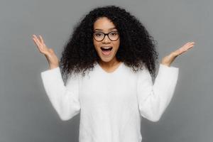 Satisfied African American woman raises hands in eureka gesture, keeps palms raised, wide opened mouth, wears optical eyewear and white jumper, isolated over grey background. Wow, its awesome. photo