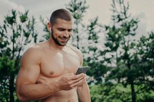 Sporty man with strong muscular body, uses smartphones and wireless earphones, downloads music for training, has thick bristle, poses outdoor against nature background. Sportsman uses modern gadget photo