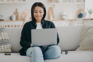 Happy girl typing on computer and smiling. Freelancer is sitting in comfortable couch with laptop. photo