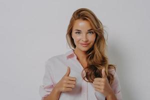 Pretty European woman with satisfied expression recommends product or best service keeps thumbs up photo