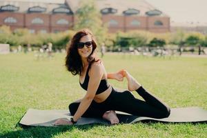 Happy fit young woman does stretching workout on fitness mat practices yoga outside dressed in activewear has strong body breathes fresh air outdoor leads active lifestyle. Sport fitness concept photo