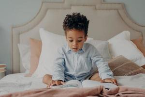 Intrigued afro american preschool child looking at colorful pictures inside of book while sitting on big bed photo