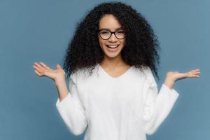 Positive friendly young woman with Afro appearance, spreads palms, holds invisible object, has charming smile, healthy skin, wears spectacles and white sweater, isolated over blue background. photo