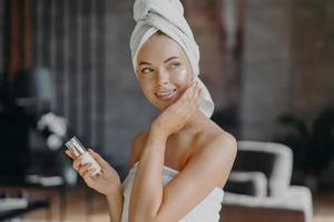 Indoor shot of young smiling woman applies moisturizer cream on face, takes care of her skin and complexion, puts lotions, has minimal makeup, wrapped in bath towel. Beauty, cosmetology concept photo