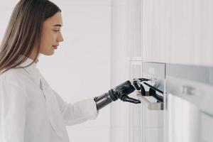 Happy disabled woman with robotic cyber hand switching oven on. Cooking at home using bionic limb. photo