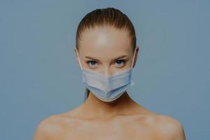 Studio shot of naked European woman wears disposable medical mask as prevention fromm coronavirus has makeup combed hair poses withh bare shoulders against blue background. Quarantine and lock down photo